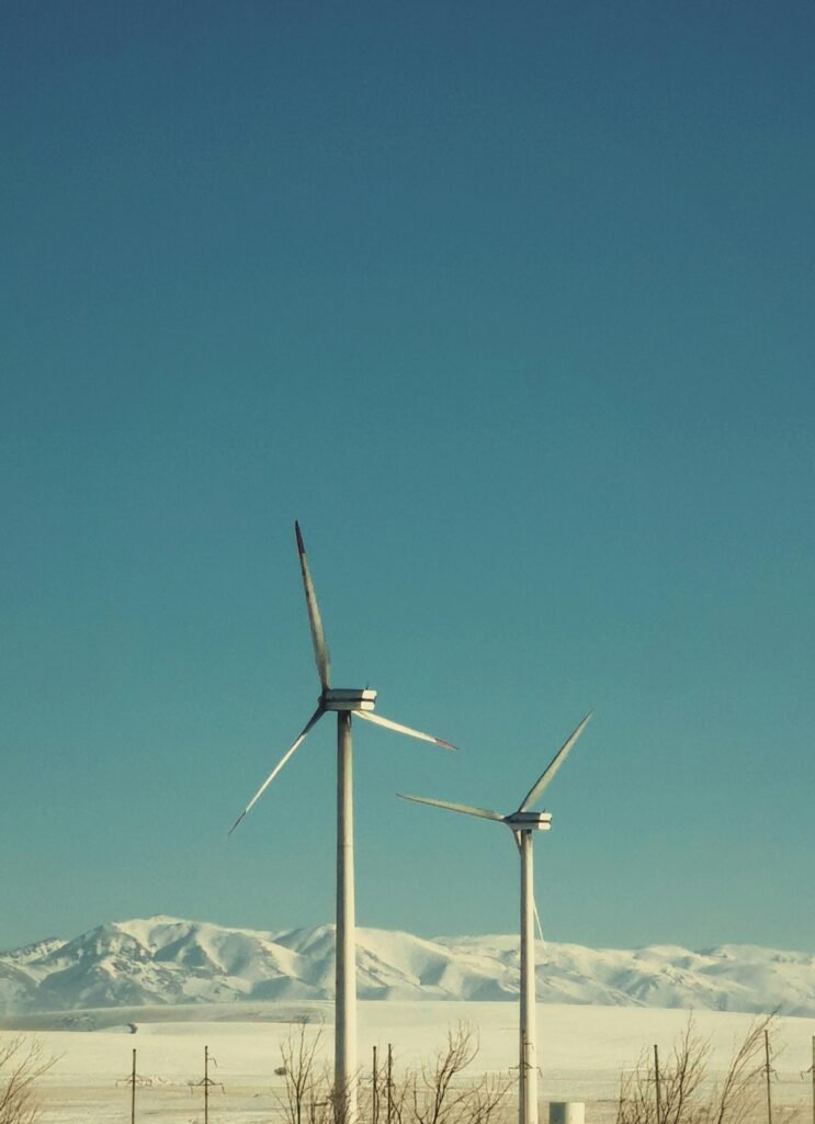 Two wind turbines stand tall against a backdrop of snow-capped mountains under a clear blue sky.